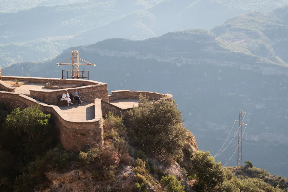 Two people sit on a stone terrace overlooking a mountain landscape, with vegetation and a structure nearby.