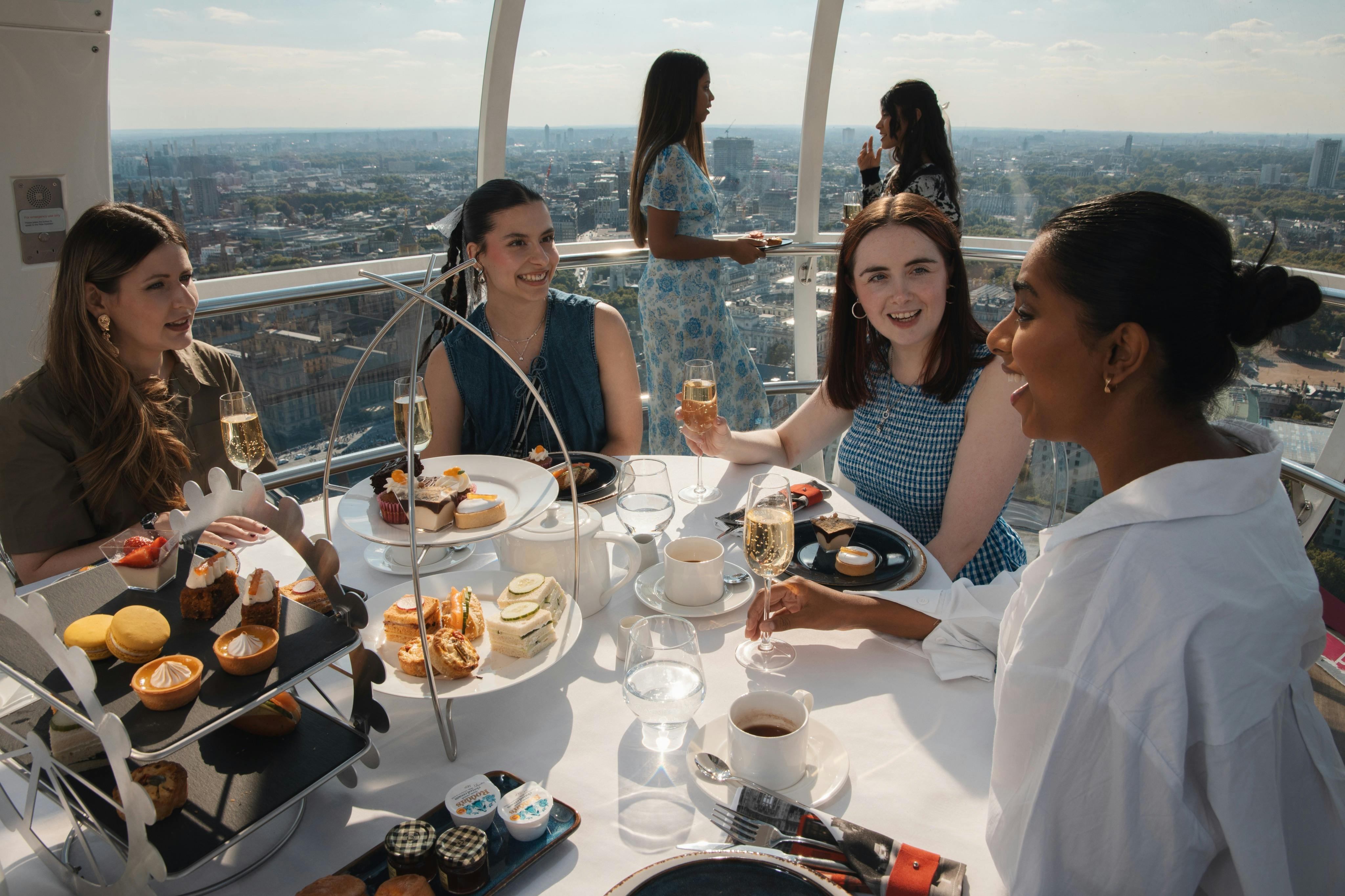 Four women enjoy a meal with drinks at a table inside a glass observation pod overlooking a cityscape.