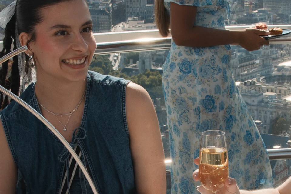 Four women enjoy a meal with drinks at a table inside a glass observation pod overlooking a cityscape.