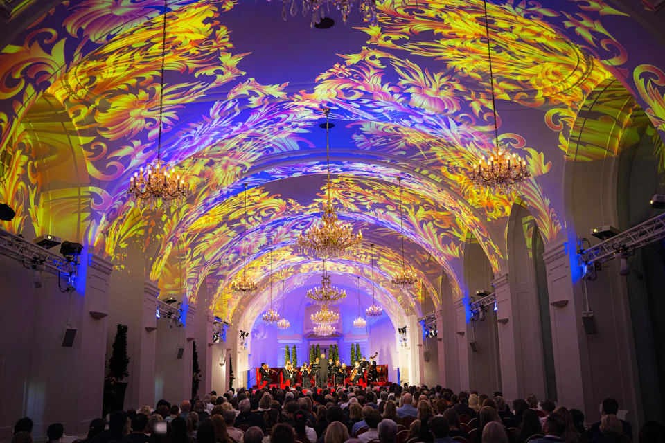 An ornate hall with decorative lighting patterns projected on the ceiling, hosting a seated audience and performers on stage.