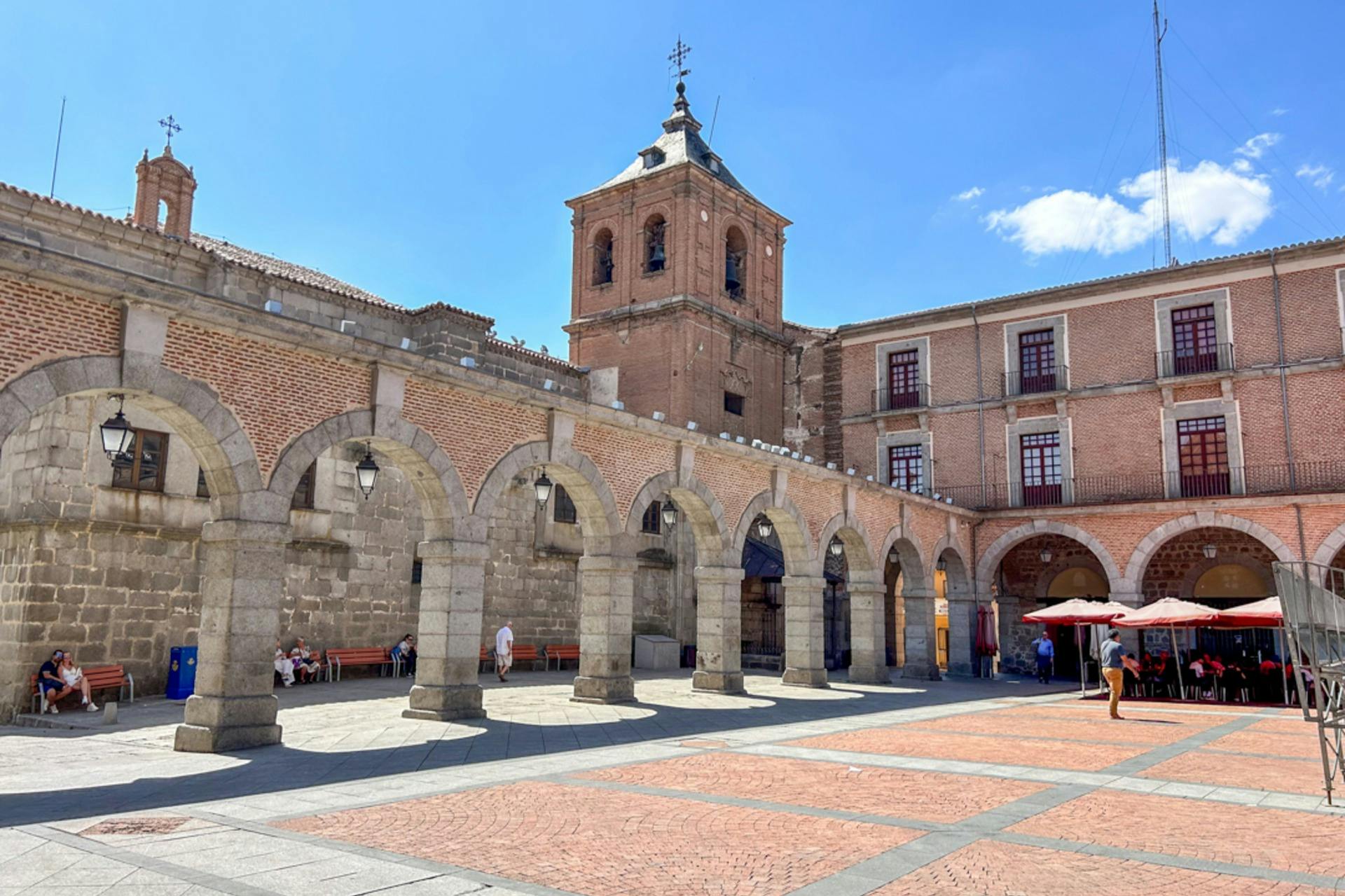 Μια αυλή με πέτρινες καμάρες, κτίρια από κόκκινα τούβλα και ένα καμπαναριό κάτω από έναν καταγάλανο ουρανό.