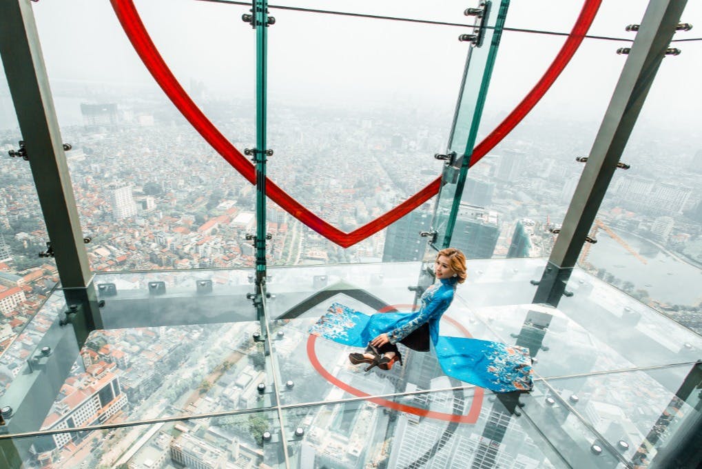 A person sits on a clear glass floor high above a city, with a red heart-shaped frame surrounding them.