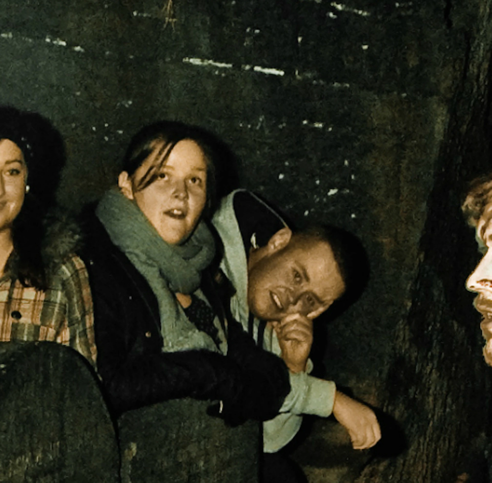 Two costumed performers interact with a group of onlookers by an old, stone wall covered in fallen leaves at night.