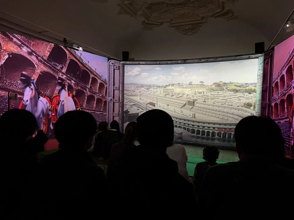 A group of people watching a projection showing historical scenes of the Roman Colosseum and an ancient city panorama.