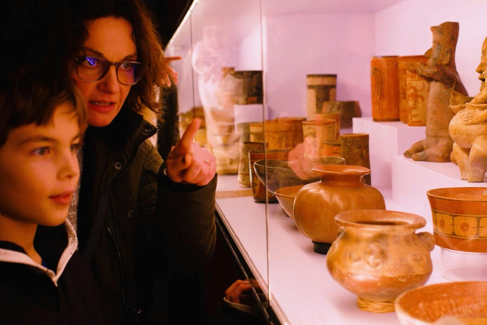 A woman and a child observe and point at artifacts displayed behind a glass case in a dimly lit museum or exhibition.