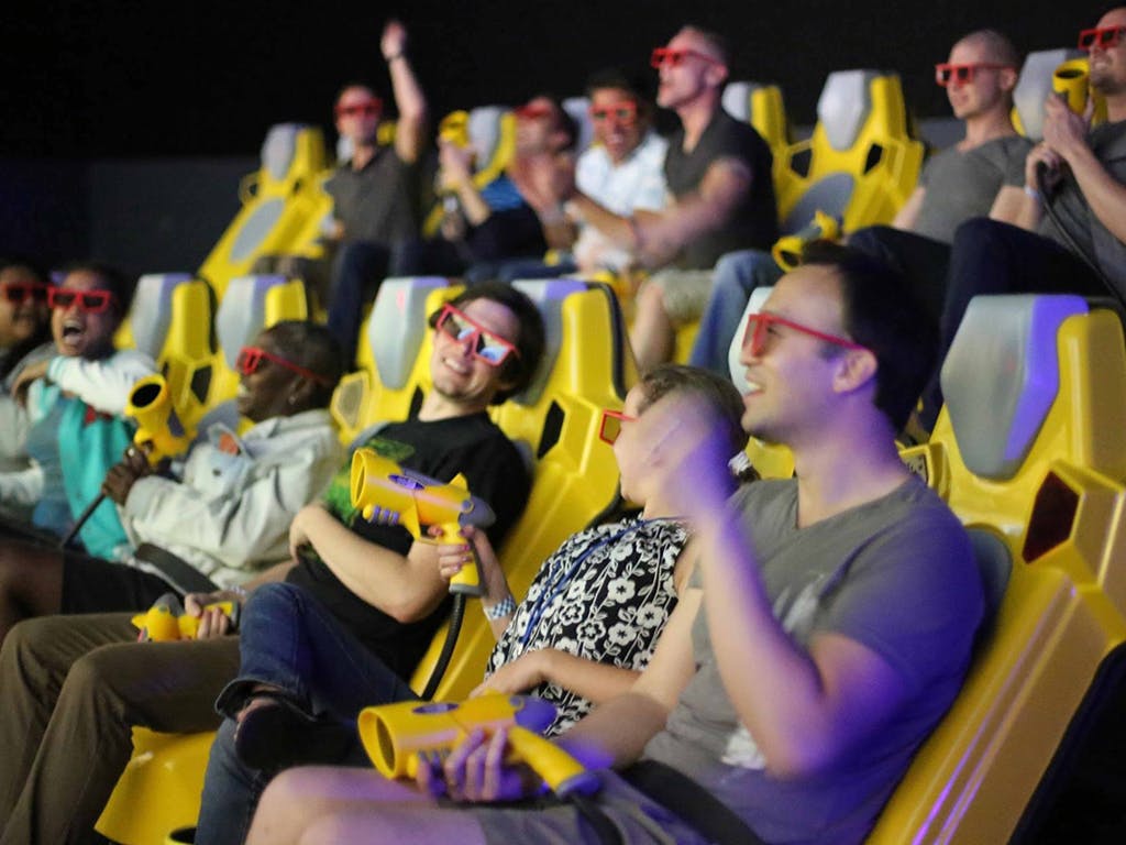 人们戴着3D眼镜，坐在黄色的椅子上，拿着玩具枪，带着一些欢呼和微笑。