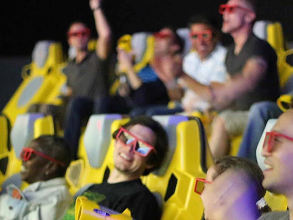 People wearing 3D glasses, seated in yellow chairs, and holding toy guns, with some cheering and smiling.