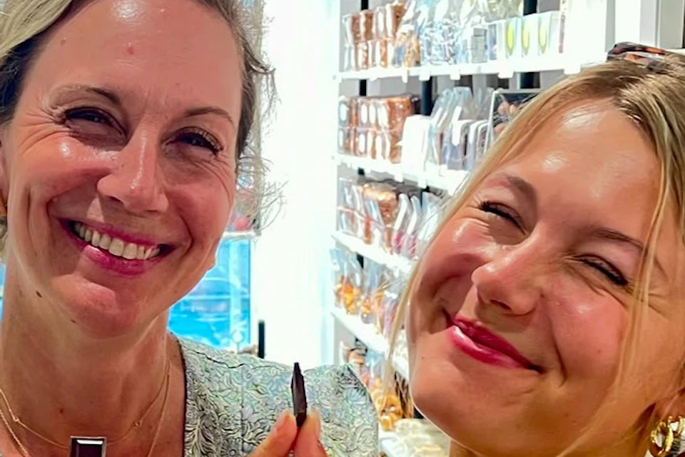 Two women smiling while holding small objects in a store with shelves of products in the background.