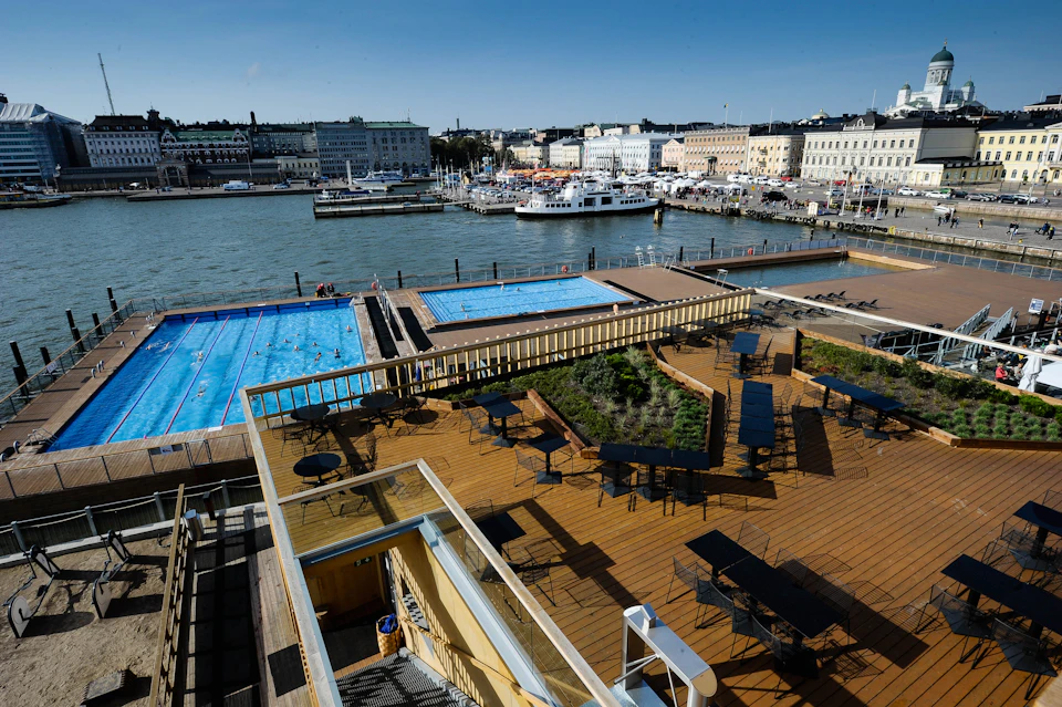Rooftop terrace with tables and chairs overlooking a harbor, featuring two swimming pools and docked boats in the background.