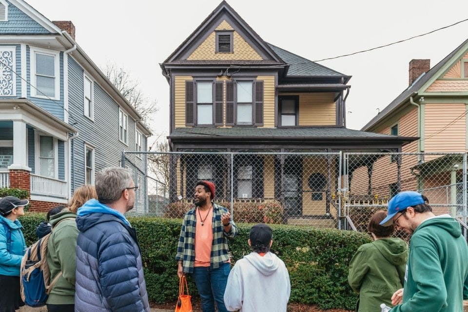 Un homme s'adresse à un petit groupe devant une maison jaune clôturée de deux étages et une maison bleue adjacente.