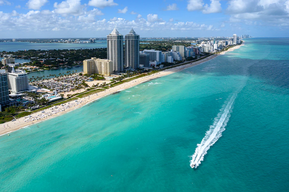Aerial view of a coastal city with high-rise buildings, a crowded beach, and a speedboat on turquoise water under a blue sky.