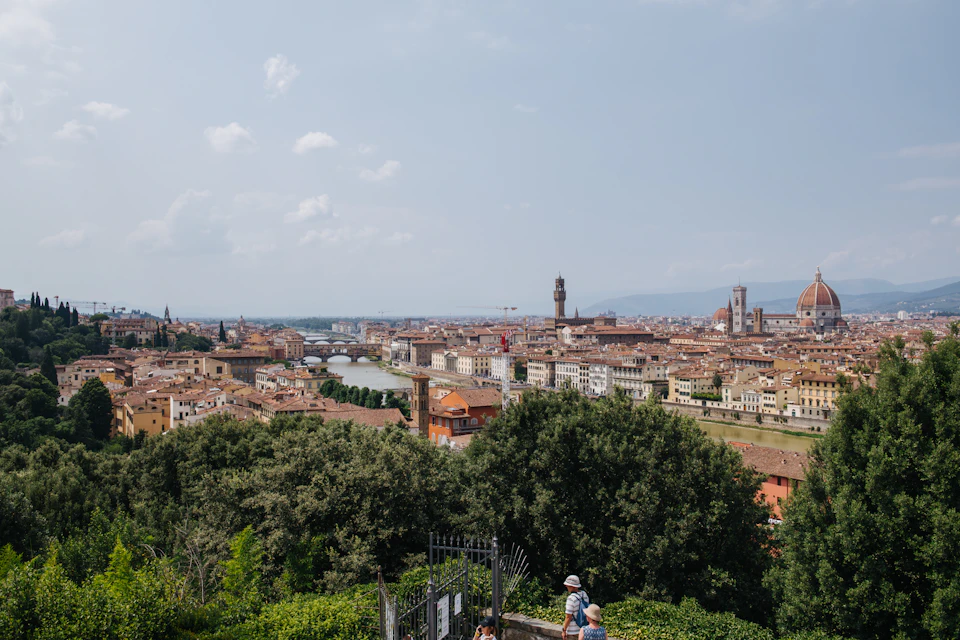 Panoramic view of a city with tall historic buildings, a river running through, and lush green trees in the foreground.