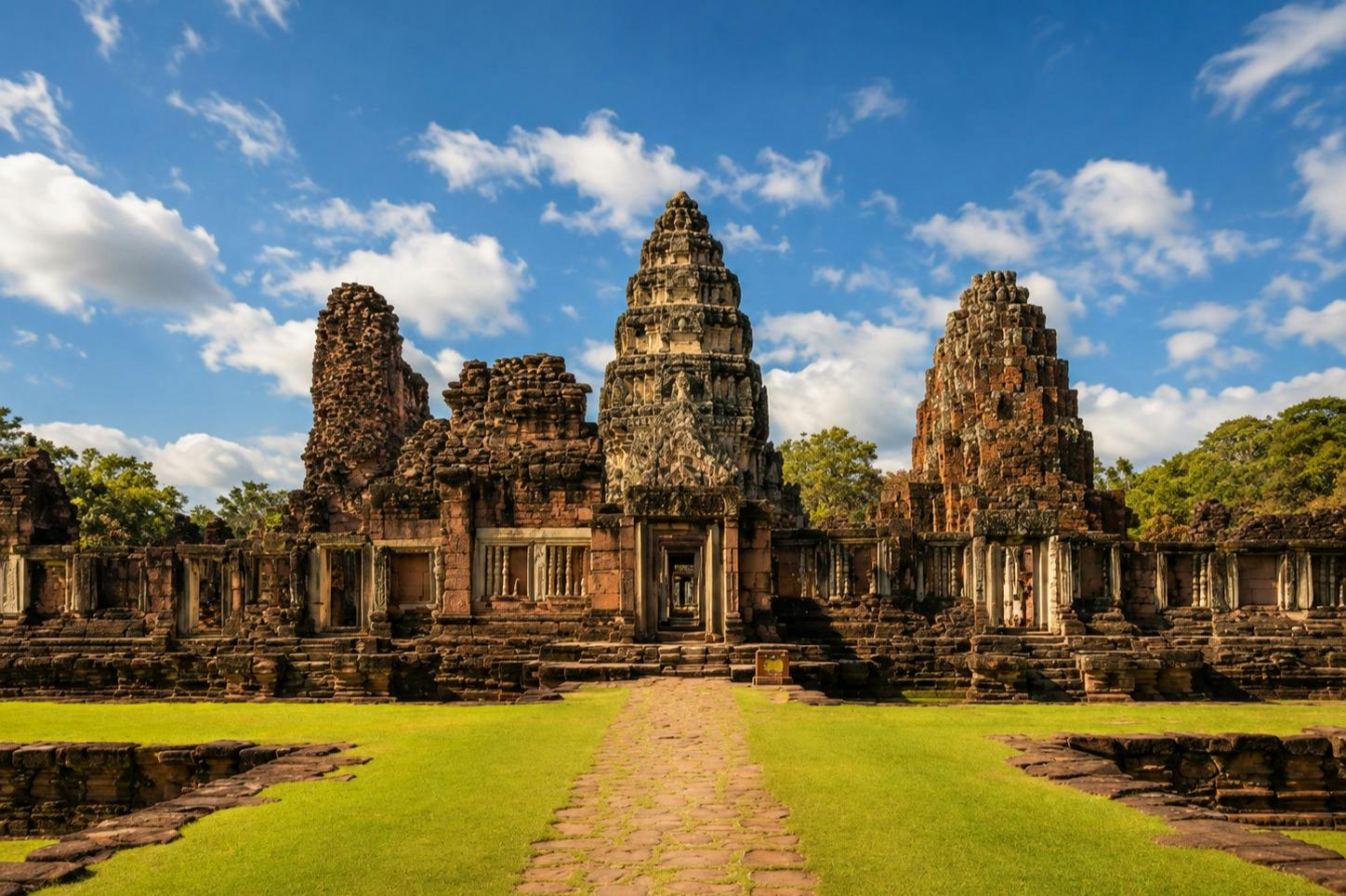Ancient stone temple ruins with intricate carvings and towers under a blue sky with clouds, surrounded by grass.