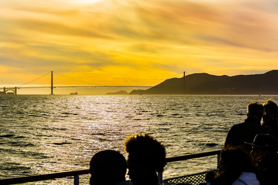 Golden Gate Bridge at sunset, viewed from a boat with silhouetted people in the foreground.