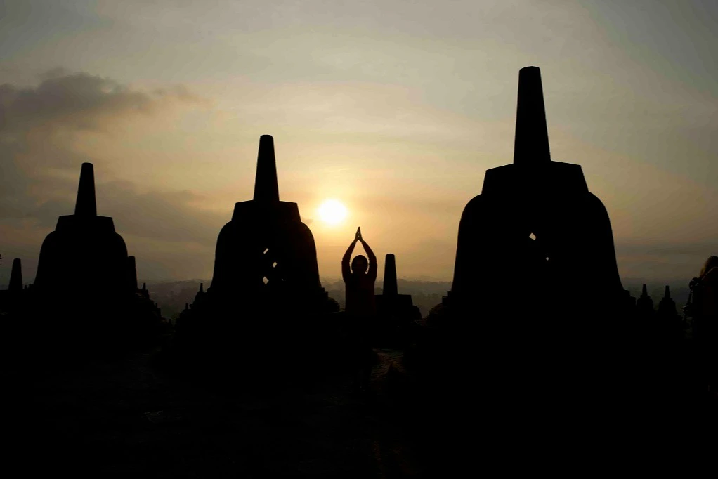 Silhouette of a person with hands raised in prayer pose at sunrise, surrounded by large bell-shaped stupas.