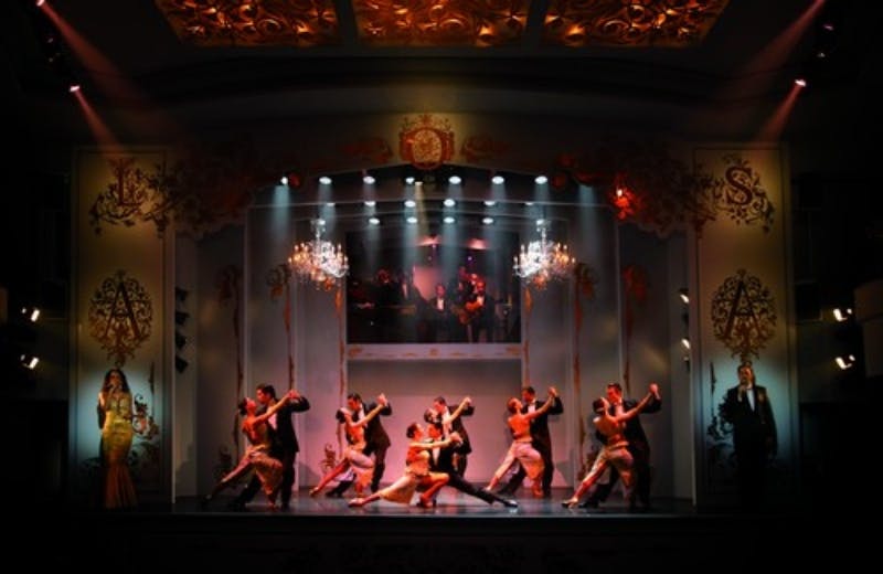 A group of dancers performs tango under chandeliers on a stage with ornate decoration and a lit orchestra in the background.
