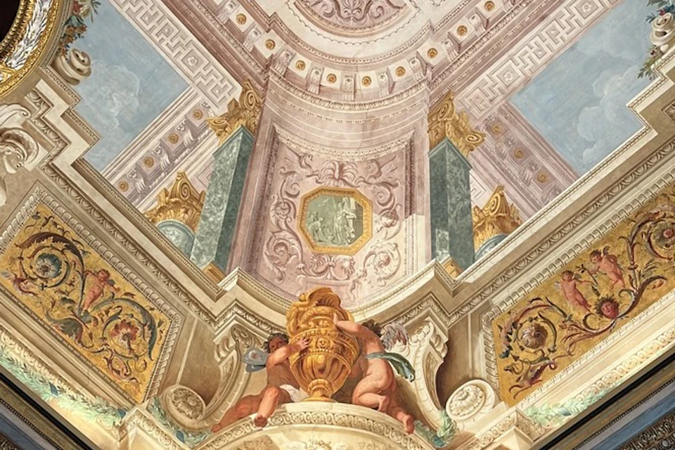 An ornate ceiling featuring classical architecture, sculptures of cherubs holding a vase, and intricate painted decorations.