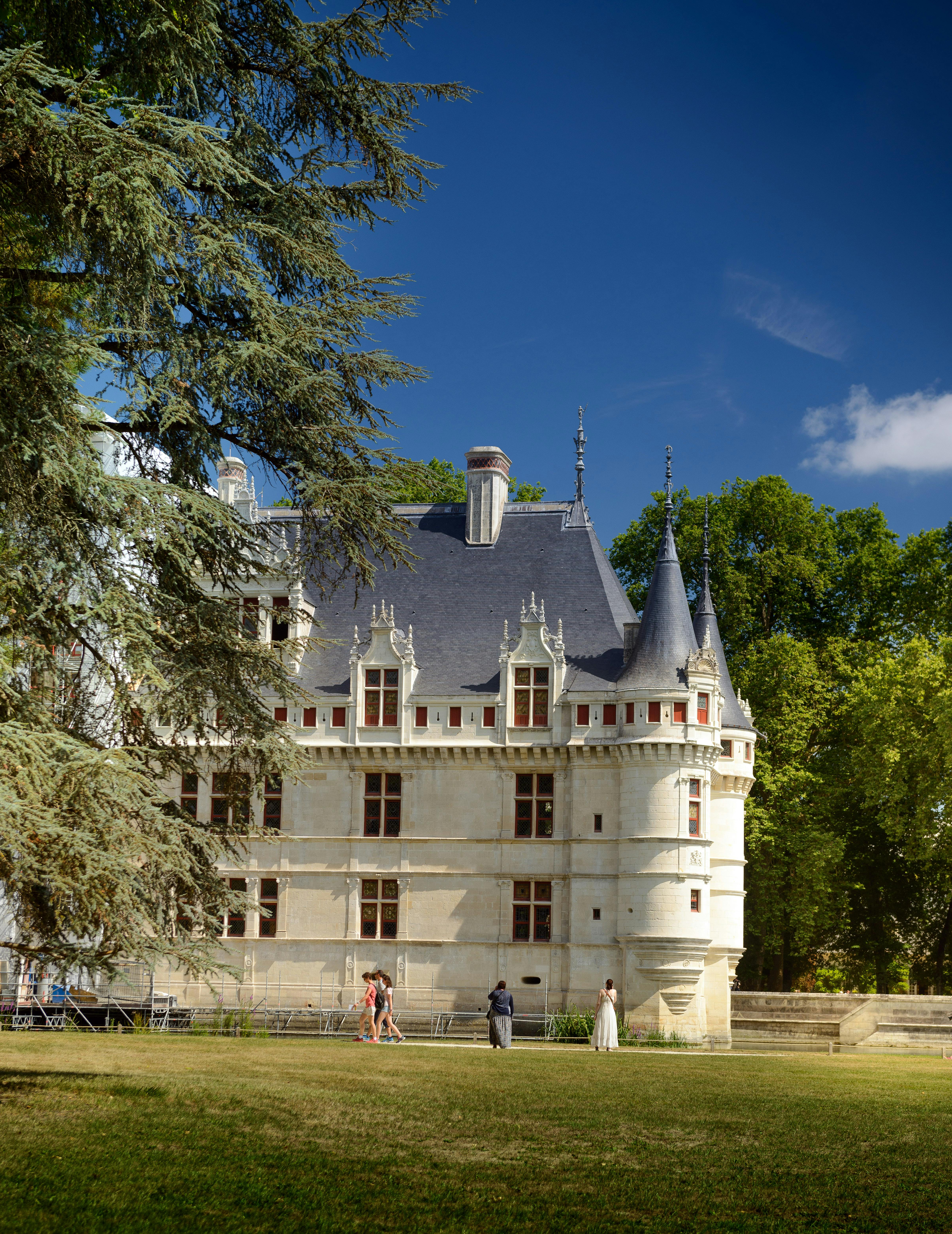 Un castello storico con torrette e finestre elaborate si erge su uno sfondo di alberi rigogliosi e un cielo azzurro e limpido. Davanti si vedono quattro persone.