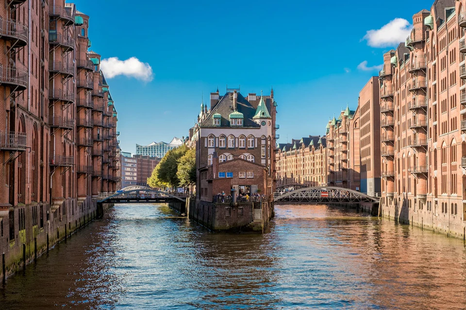 A historic building with green roofs stands between canal waters, flanked by brick buildings and bridges under a clear blue sky.