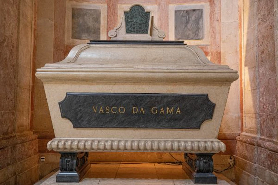A stone tomb prominently inscribed with "Vasco Da Gama," featuring ornate details and set against a backdrop of marbled walls.