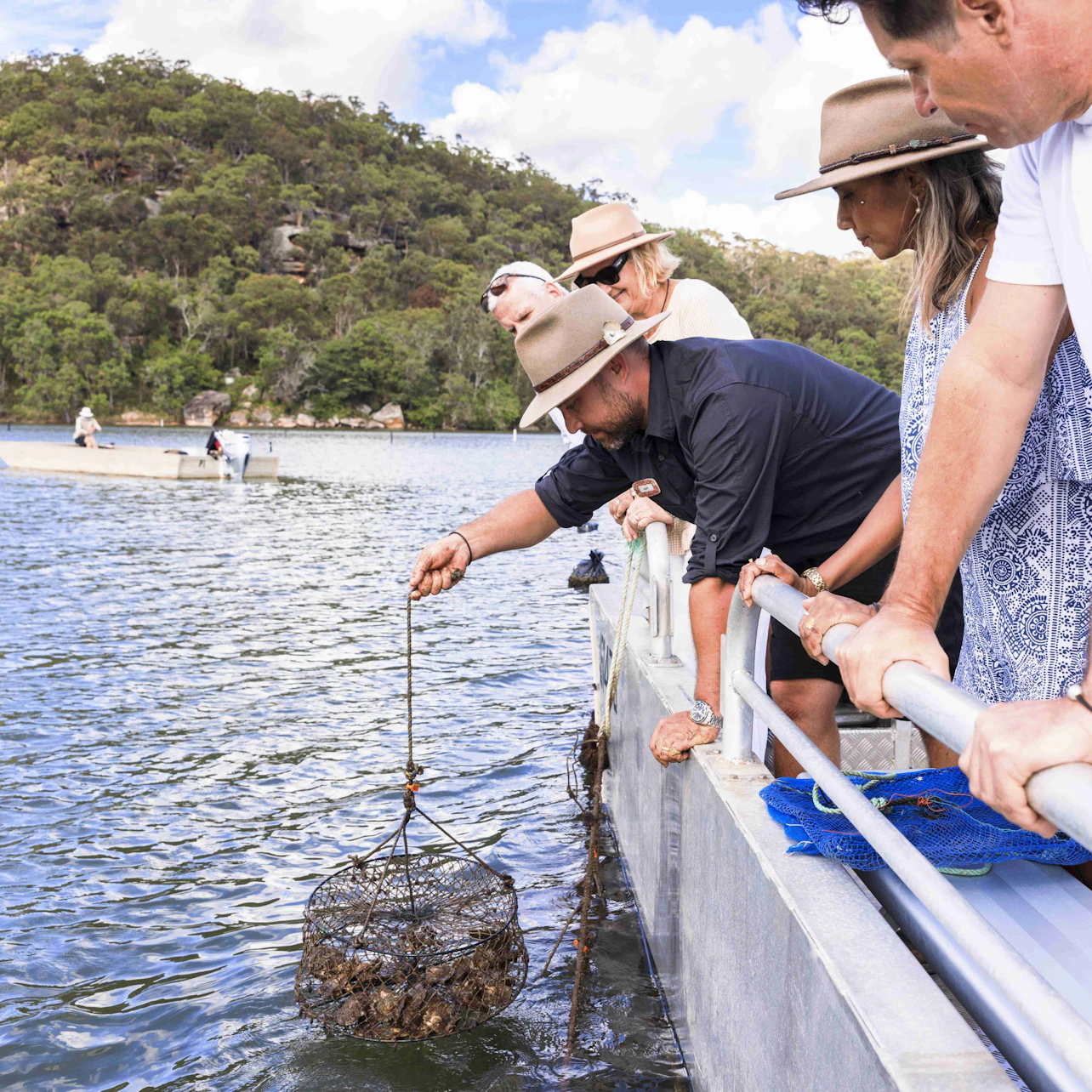 A group of people lean over a railing on a boat, observing a net basket being pulled up from the water in a scenic lake area.