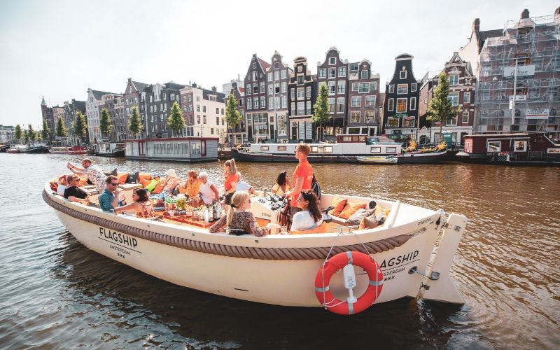 People on a tour boat in an Amsterdam canal, with historic buildings and houseboats in the background.