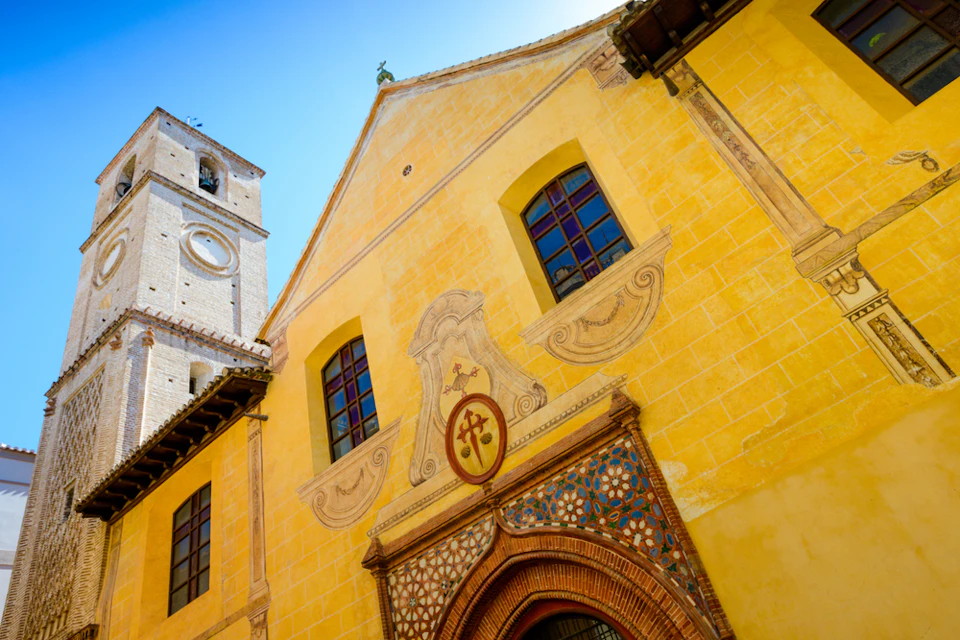 Yellow stucco building with ornate archway, two large windows, and a decorative crest. Adjacent stone tower on the left. Clear blue sky.