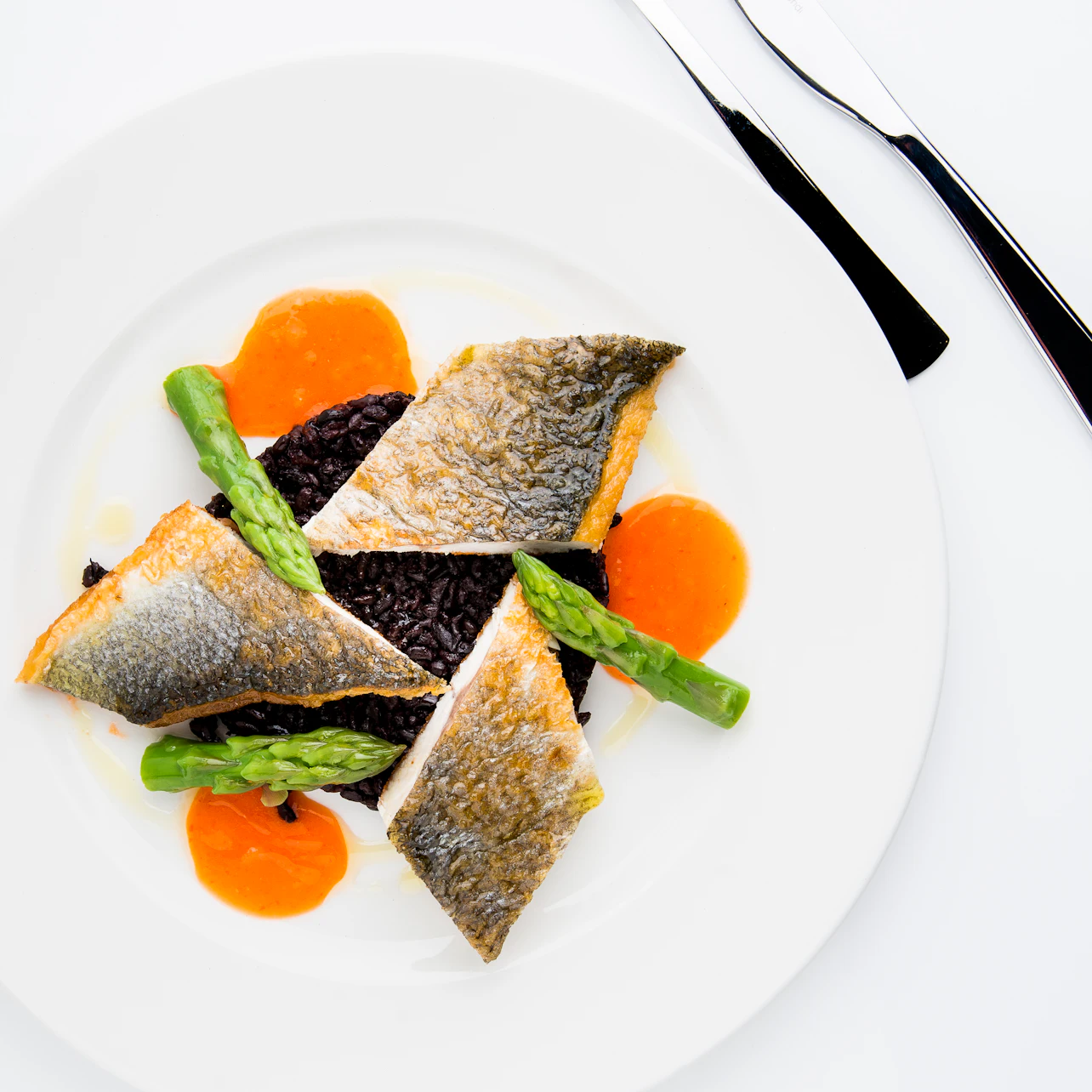 A gourmet dish featuring seared fish fillets, black rice, asparagus, and orange sauce, elegantly plated on a white dish with cutlery.