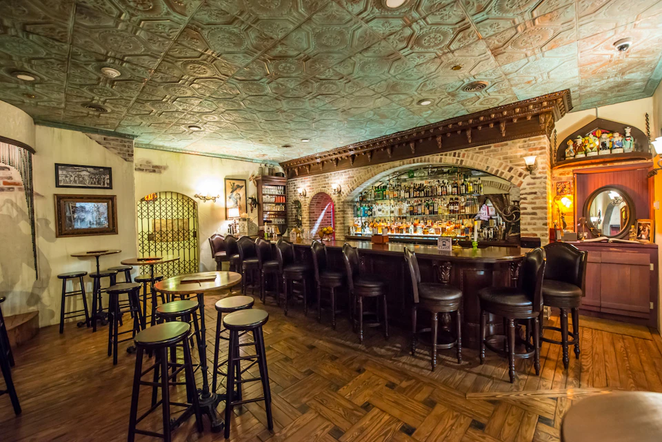 A cozy, dimly lit bar with a vintage decor, featuring a wooden bar counter, cushioned bar stools, and shelves filled with bottles.