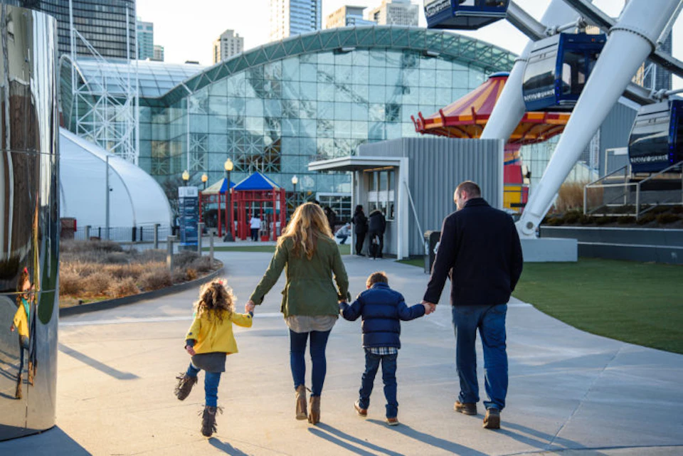 A family of four walking hand in hand towards a large glass building, with colorful structures and people in the background.