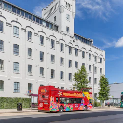 A red open-top double-decker tour bus in front of a large, white building on a sunny day.