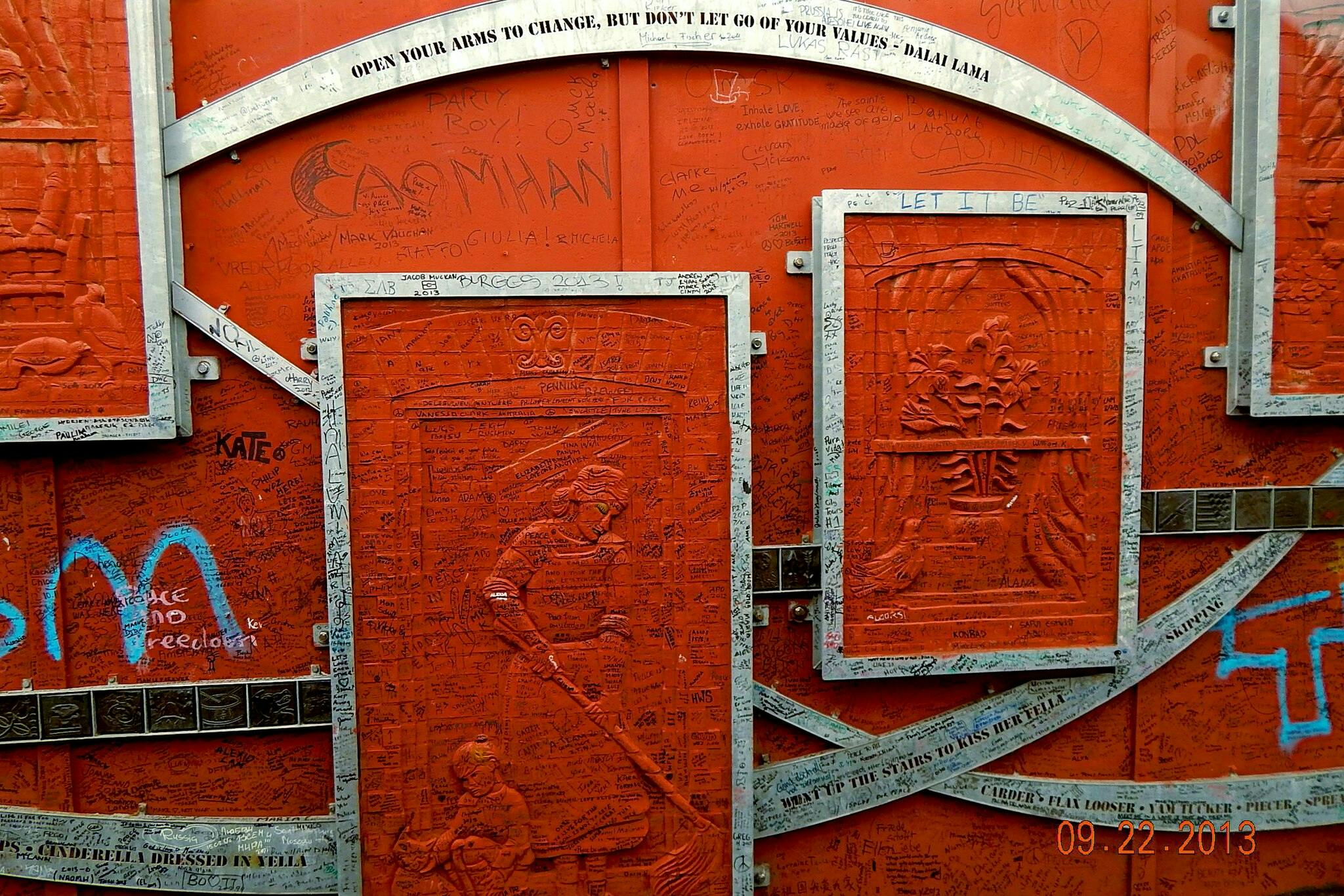 Červená zeď pokrytá různými podpisy a zprávami. Kovové rámy zvýrazňují gravírované panely s citátem připisovaným dalajlámovi výše.
