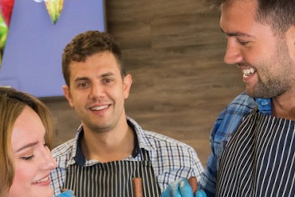 Group of people in striped aprons smiling and stirring chocolate in pitchers in a kitchen setting.