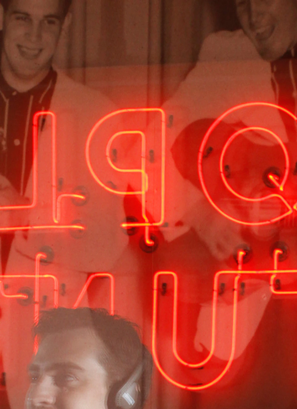 A young man with headphones stands in front of a neon sign and a photo of musicians performing.