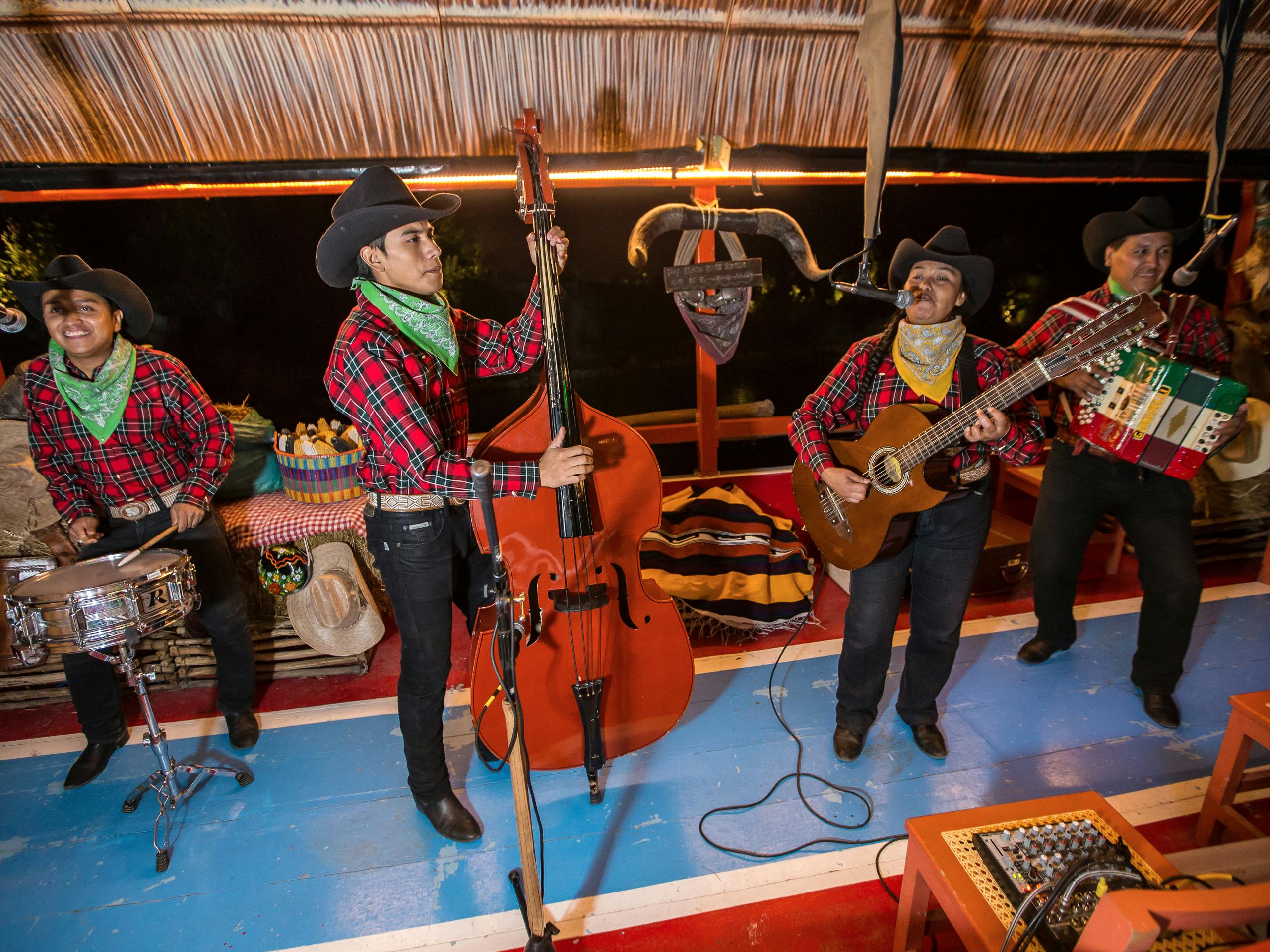 Τρεις μουσικοί με καουμπόικα καπέλα και καρό πουκάμισα παίζουν με διπλό μπάσο, κιθάρα και τύμπανα σε μια πολύχρωμη σκηνή.