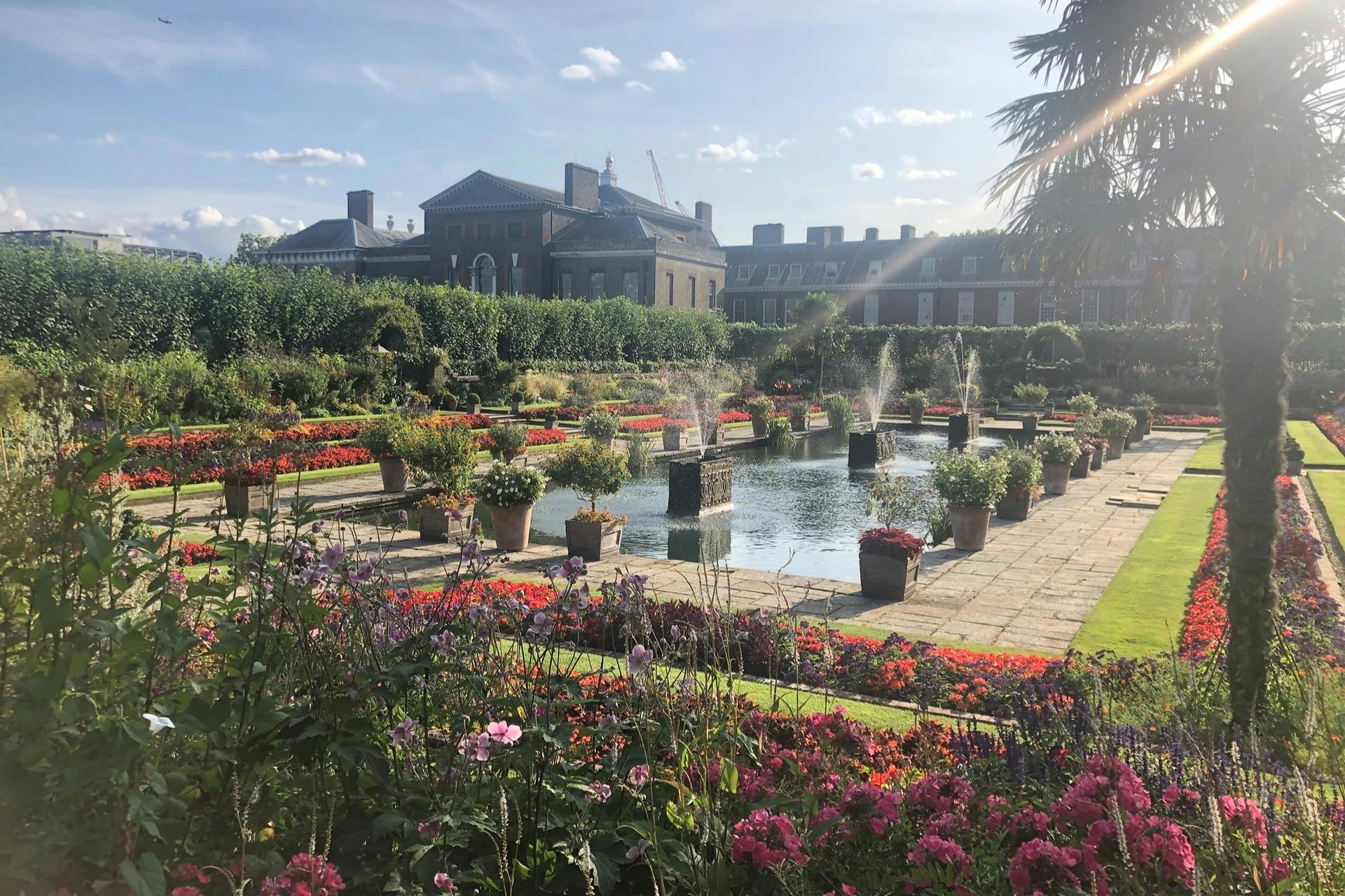 Formal garden with colorful flowerbeds, a rectangular pond with fountains, and a large historic building in the background.