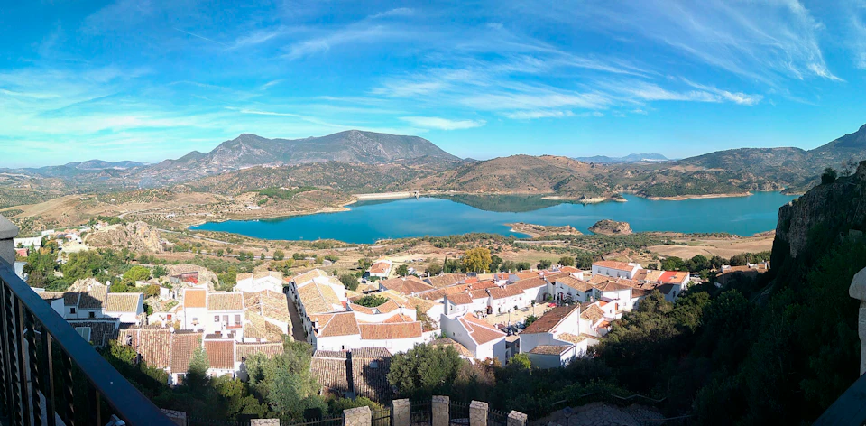 A scenic view of a village with white-roofed houses next to a lake, surrounded by hills and under a blue sky with light clouds.