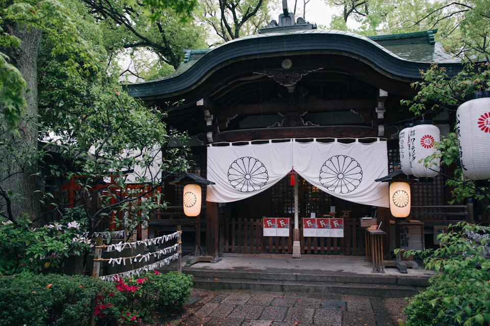 A traditional Japanese shrine entrance with white curtains, paper lanterns, and nearby greenery.