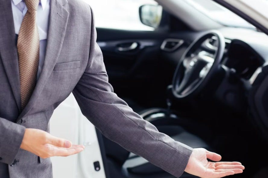 A person in a gray suit extends their arm towards an open car door, inviting entry.