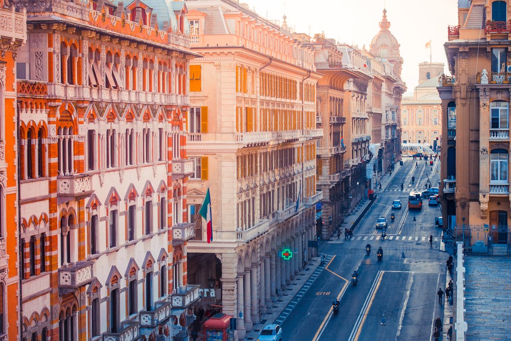 Ένας δρόμος της πόλης με περίτεχνα, ψηλά κτίρια, μοτοσικλέτες και λίγα αυτοκίνητα στο δρόμο, και ορατή μια ιταλική σημαία.