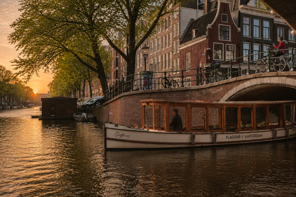 A canal boat passing under a bridge in a city with historic buildings and trees during sunset.