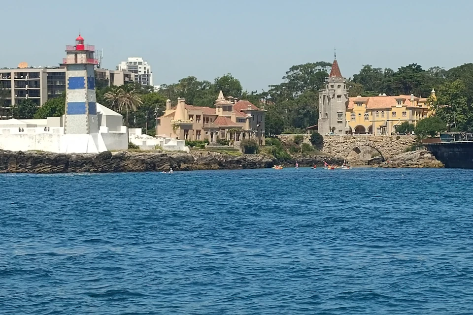 Coastal scene with a lighthouse, historic buildings, and a rocky shoreline. A few people are kayaking on the vibrant blue water.
