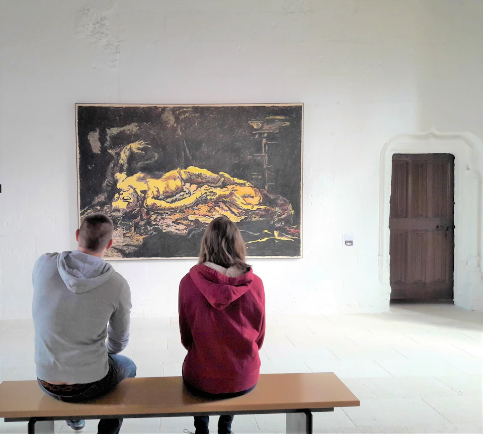 Two people sit on a bench, viewing a large painting in a museum room with white walls and a wooden door.