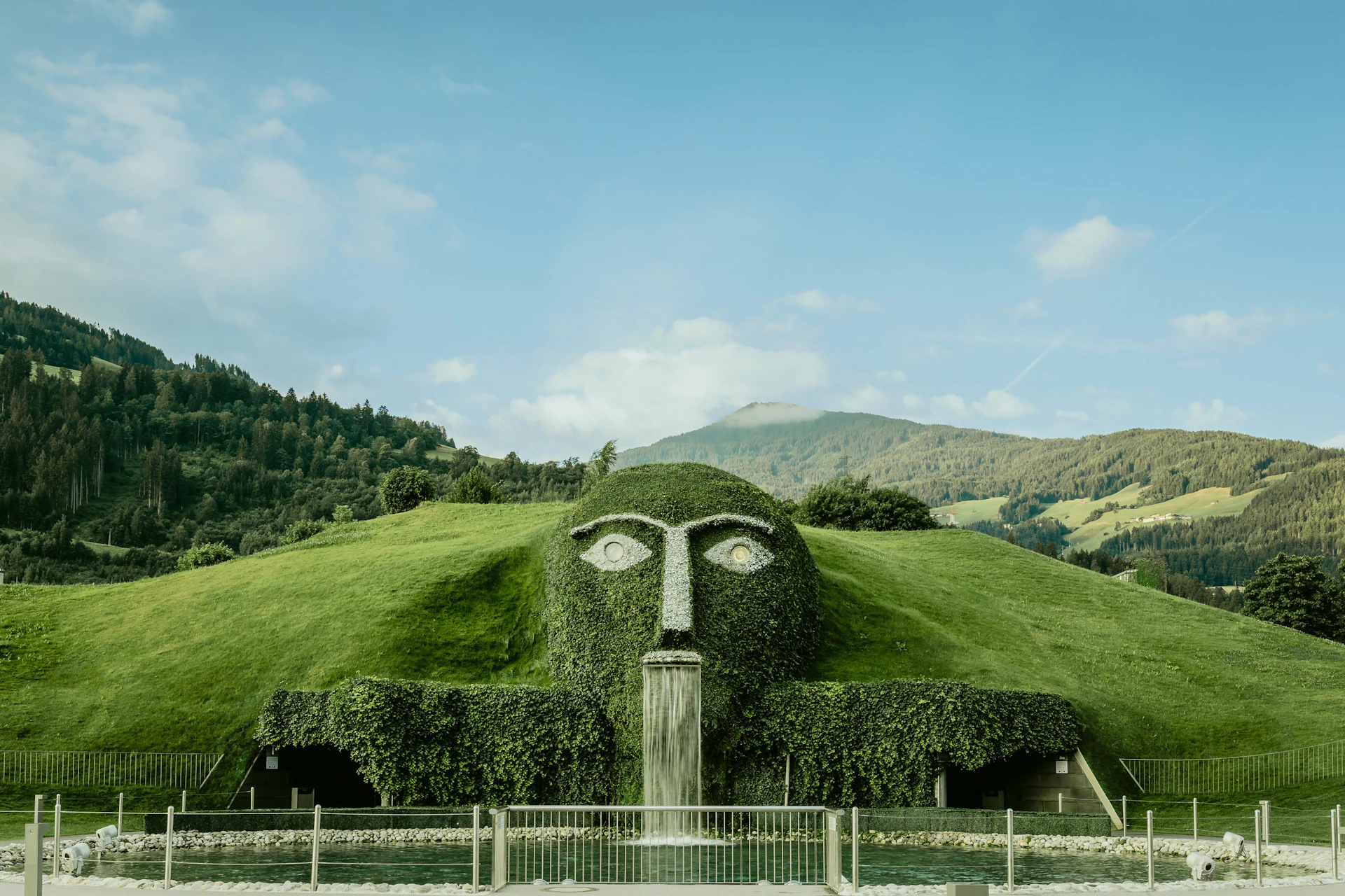 A hillside sculpture of a face with a waterfall mouth, surrounded by greenery and set against a mountainous backdrop.