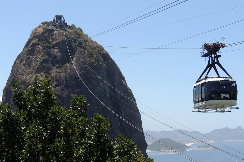 A cable car ascending a mountain with a station at the top; lush greenery and ocean visible in the background.