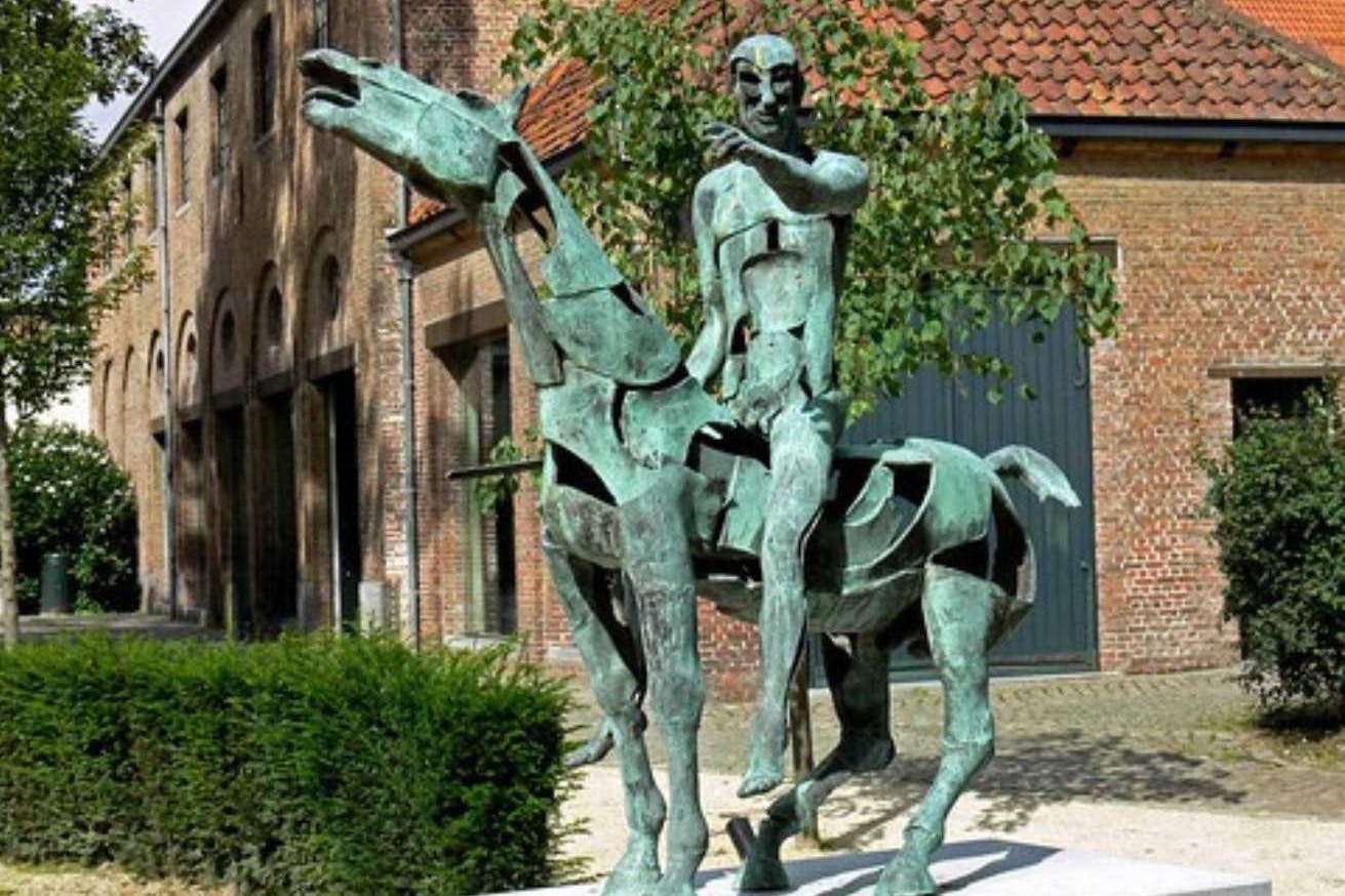 一尊骑马男子的青铜雕像，坐落在室外区域，背景是砖砌建筑和绿化植物。