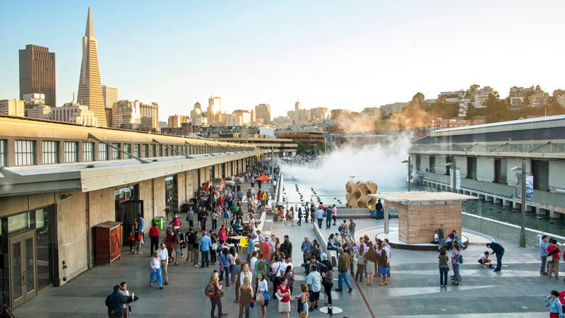 도시의 스카이라인을 배경으로 현대적인 건물과 수로에서 솟아오른 안개가 펼쳐진 탁 트인 광장에 많은 사람들이 모여듭니다.