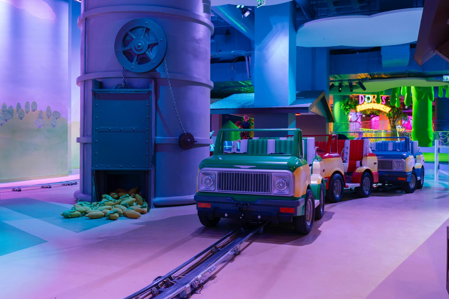 Een kleurrijke indoorattractie met een groene tractor op een baan bij een silo en maïsdecor onder levendige verlichting.