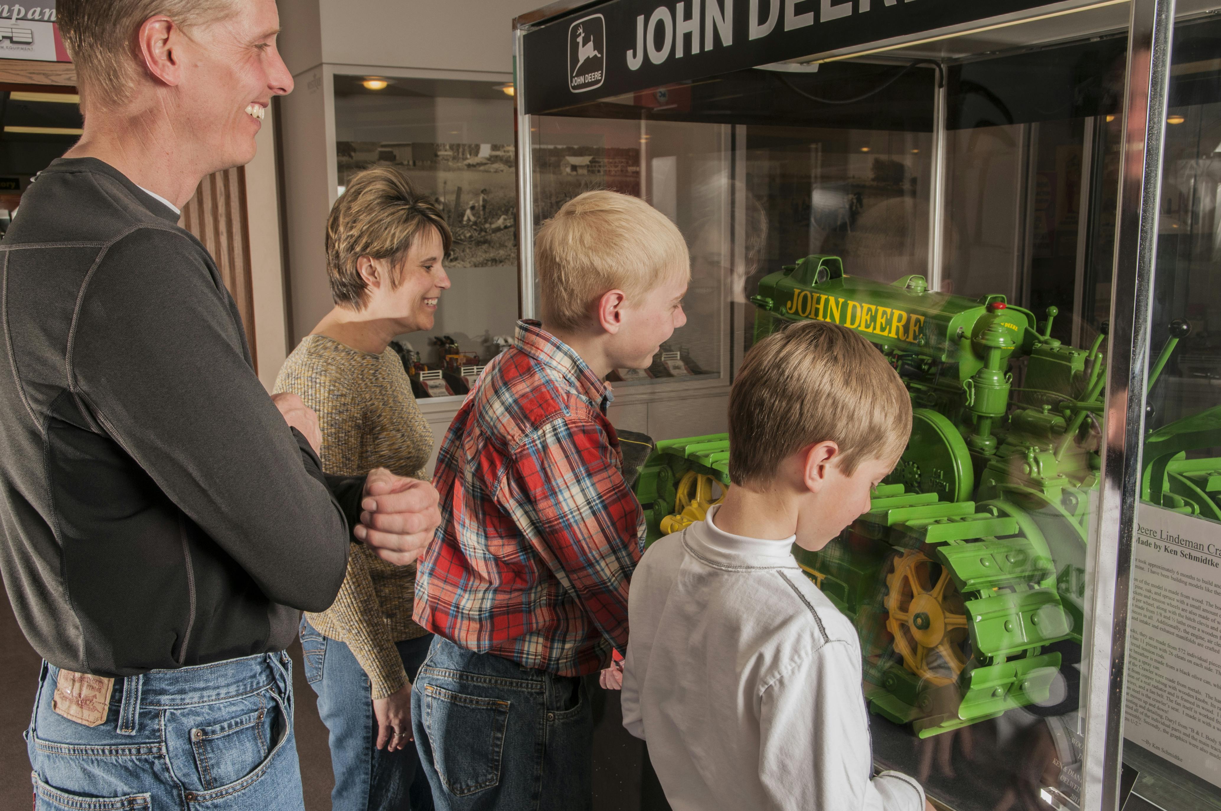 Μια τετραμελής οικογένεια παρατηρεί μια οθόνη John Deere, με δύο αγόρια να επιθεωρούν προσεκτικά ένα πράσινο και κίτρινο μοντέλο τρακτέρ.