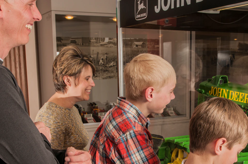 A family of four observes a John Deere display, with two boys closely inspecting a green and yellow model tractor.